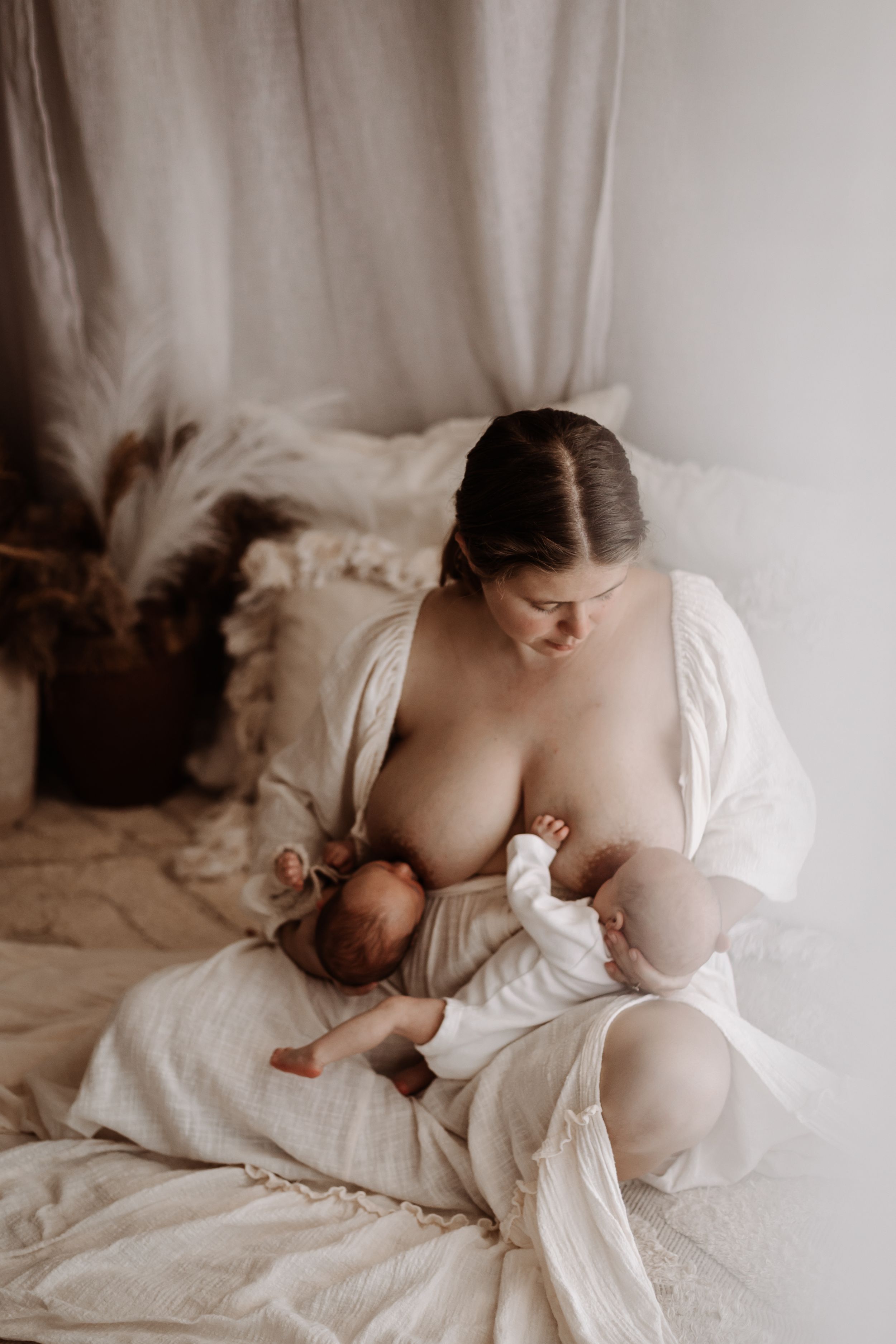 A mother in a white robe tenderly breastfeeding her newborn baby in a soft, neutral-toned studio setting.