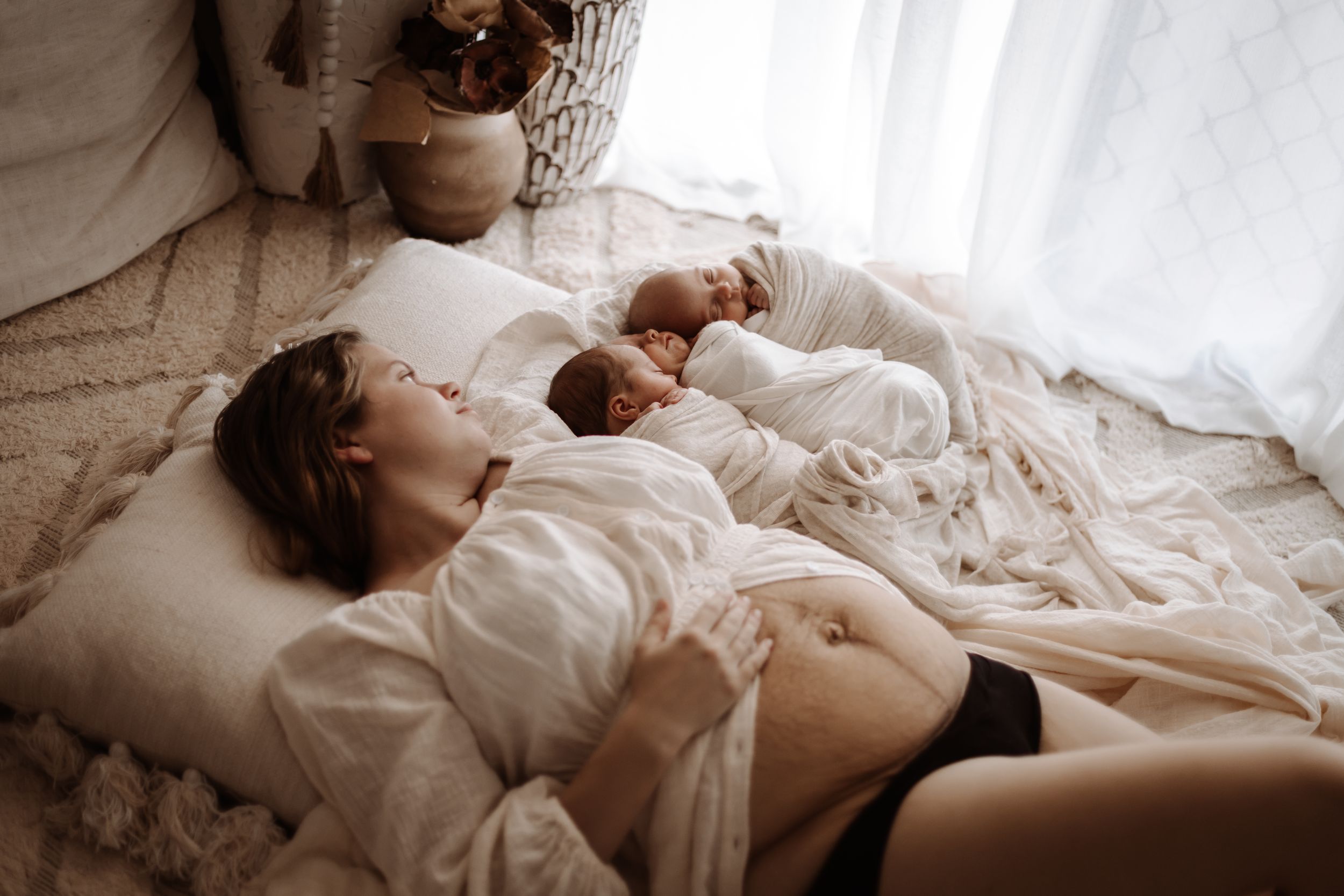 Mother resting on bed with newborn twins in soft natural light, postpartum lifestyle photography.