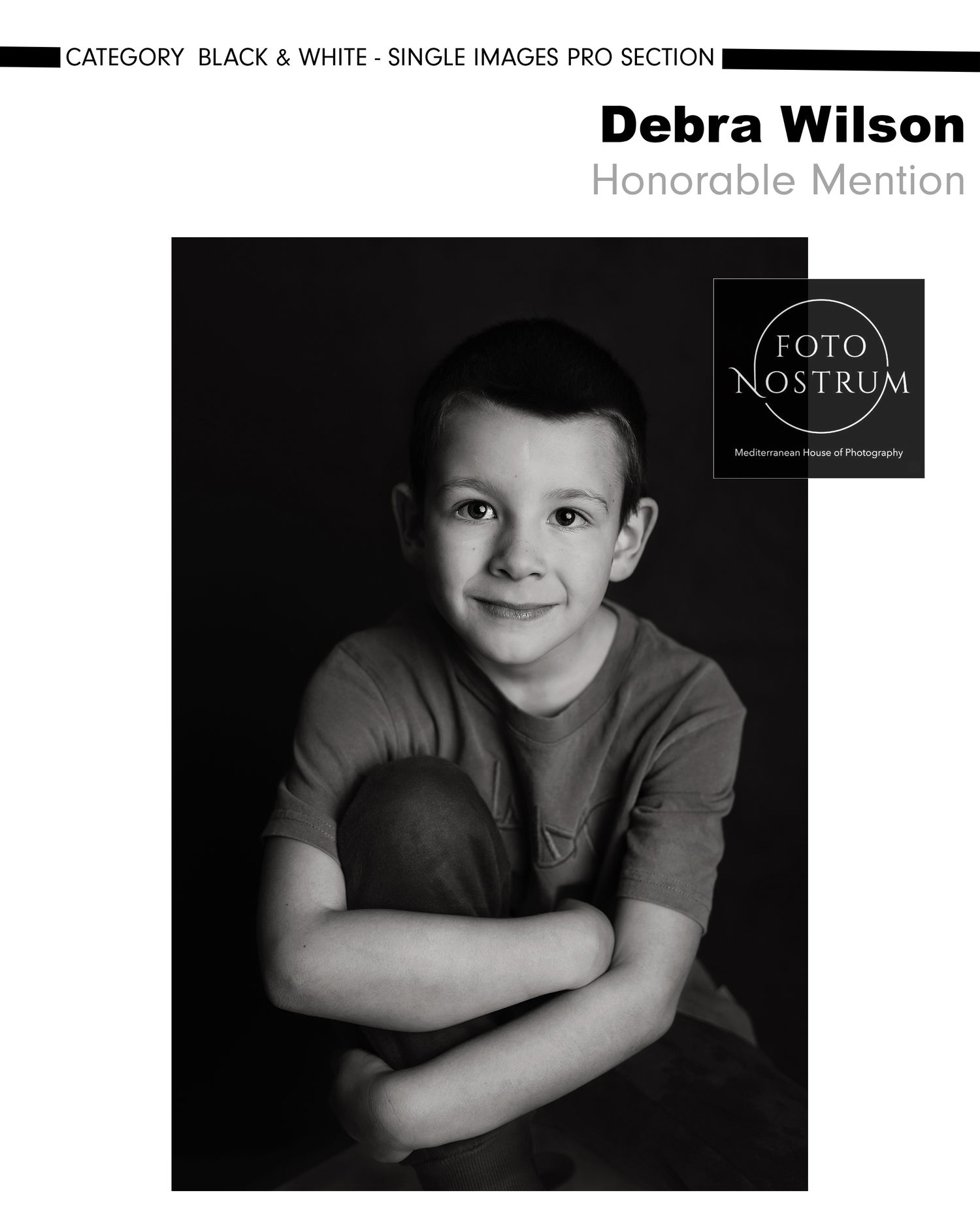Black and white portrait of a young boy hugging a ball, smiling softly, by Debra Wilson, Honorable Mention.
