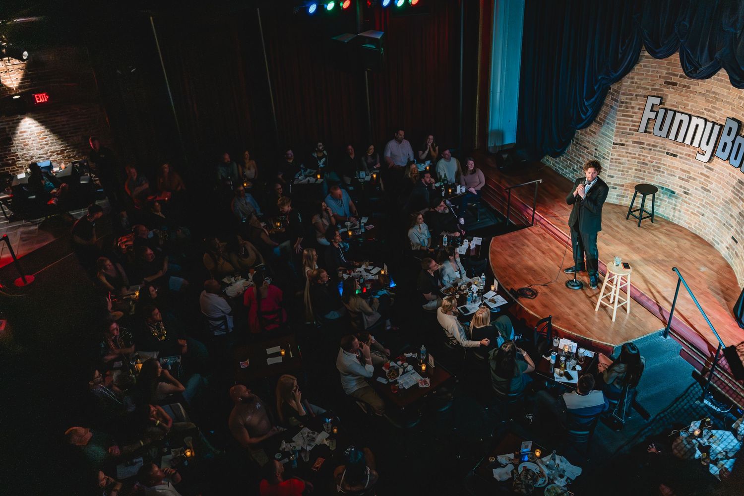 A male stand-up comedian performs on stage at a comedy club, holding a microphone in front of a brick wall backdrop.