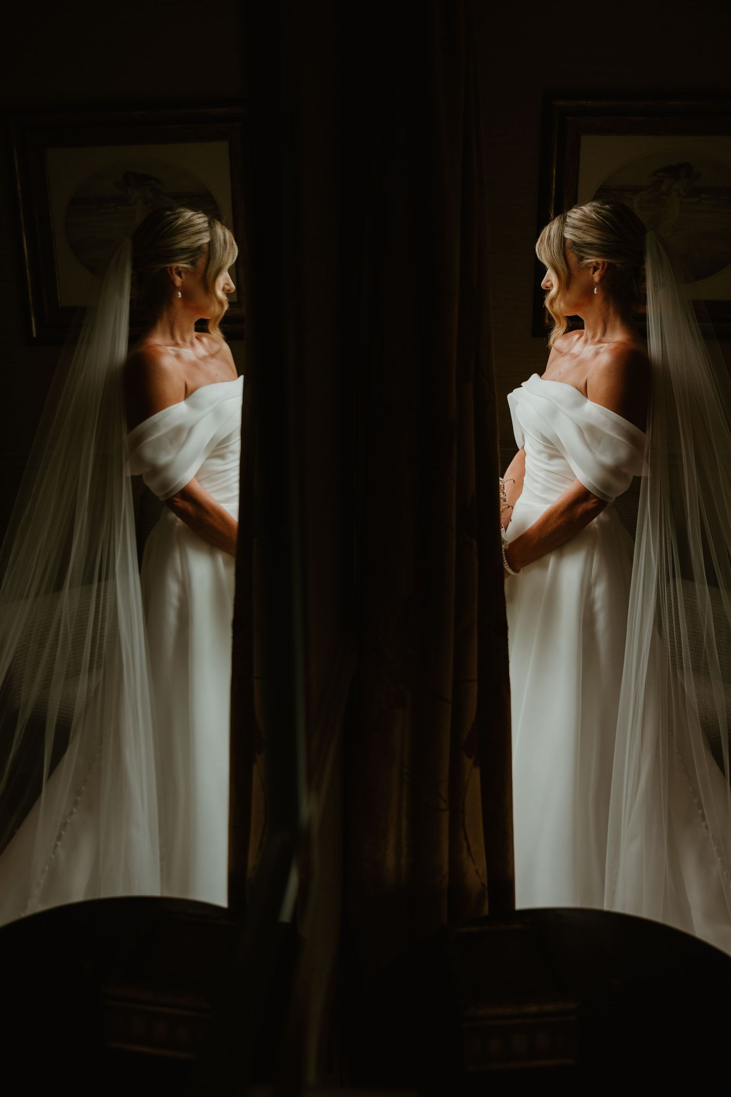 A bride in a white off-shoulder gown and veil stands in a dark room, reflected symmetrically in a mirror.