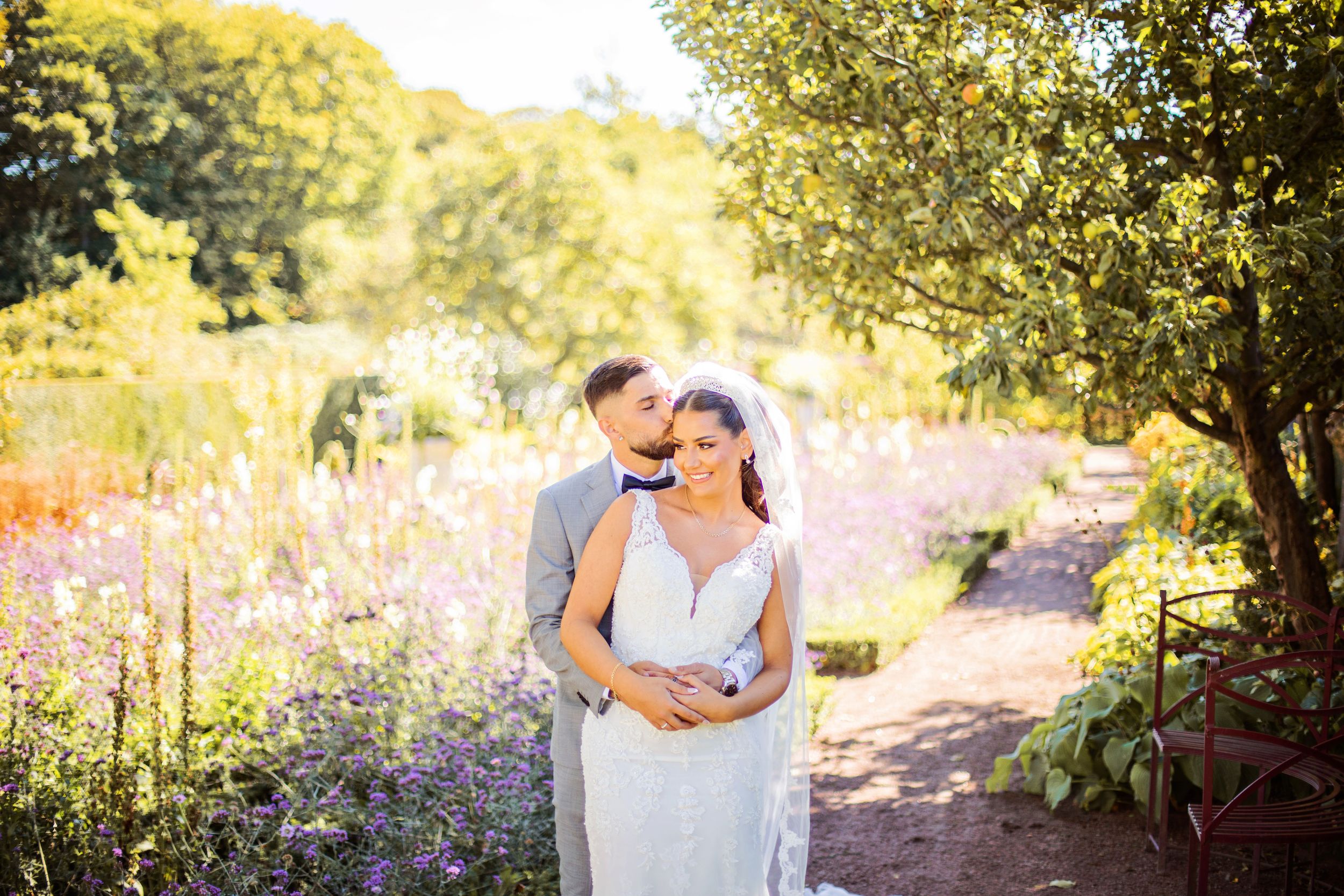 Bride and groom embrace in a sunlit garden with colorful wildflowers and lush greenery on their wedding day.