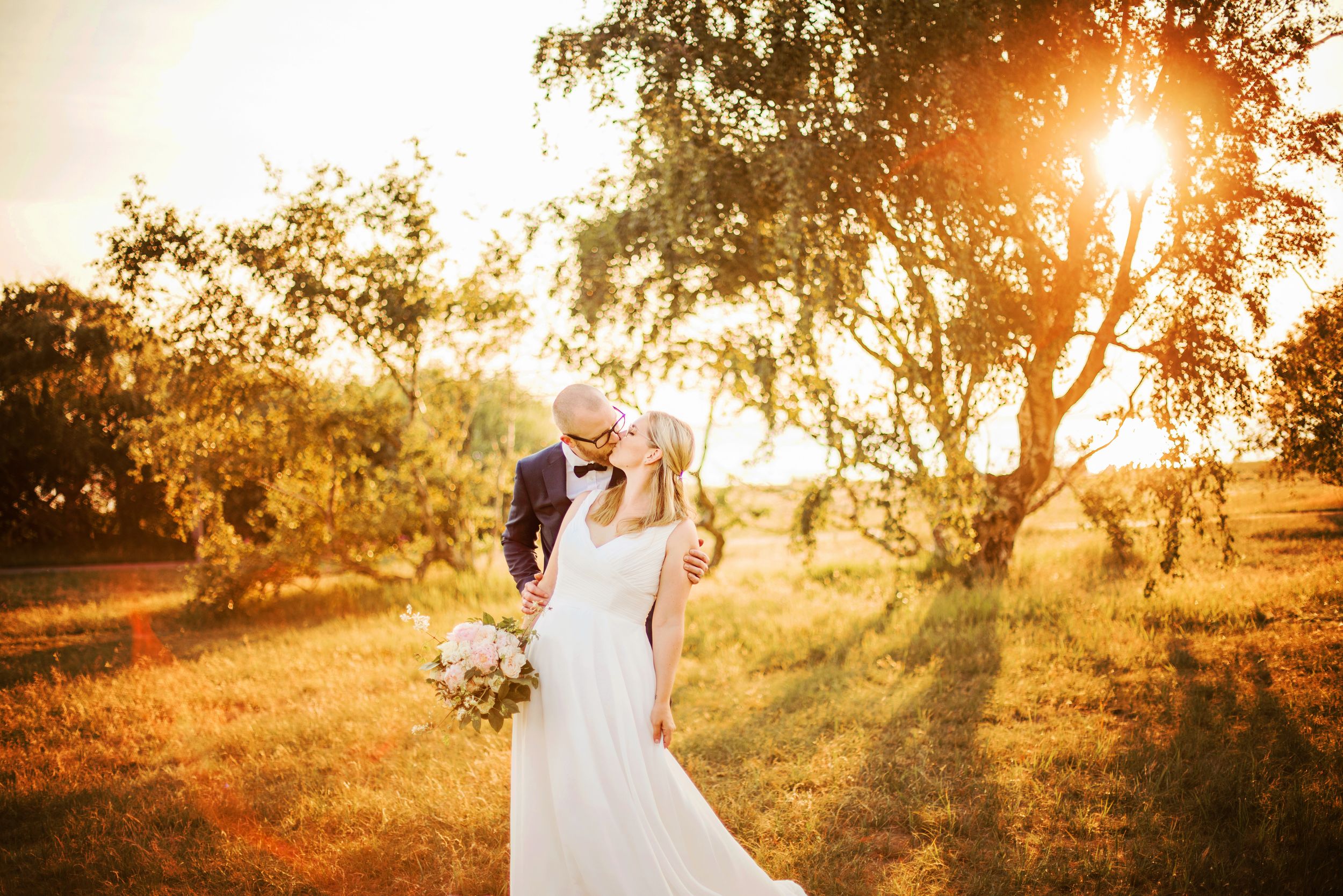 Bride and groom embracing in golden sunset light among trees, bride holding bouquet in white wedding dress.