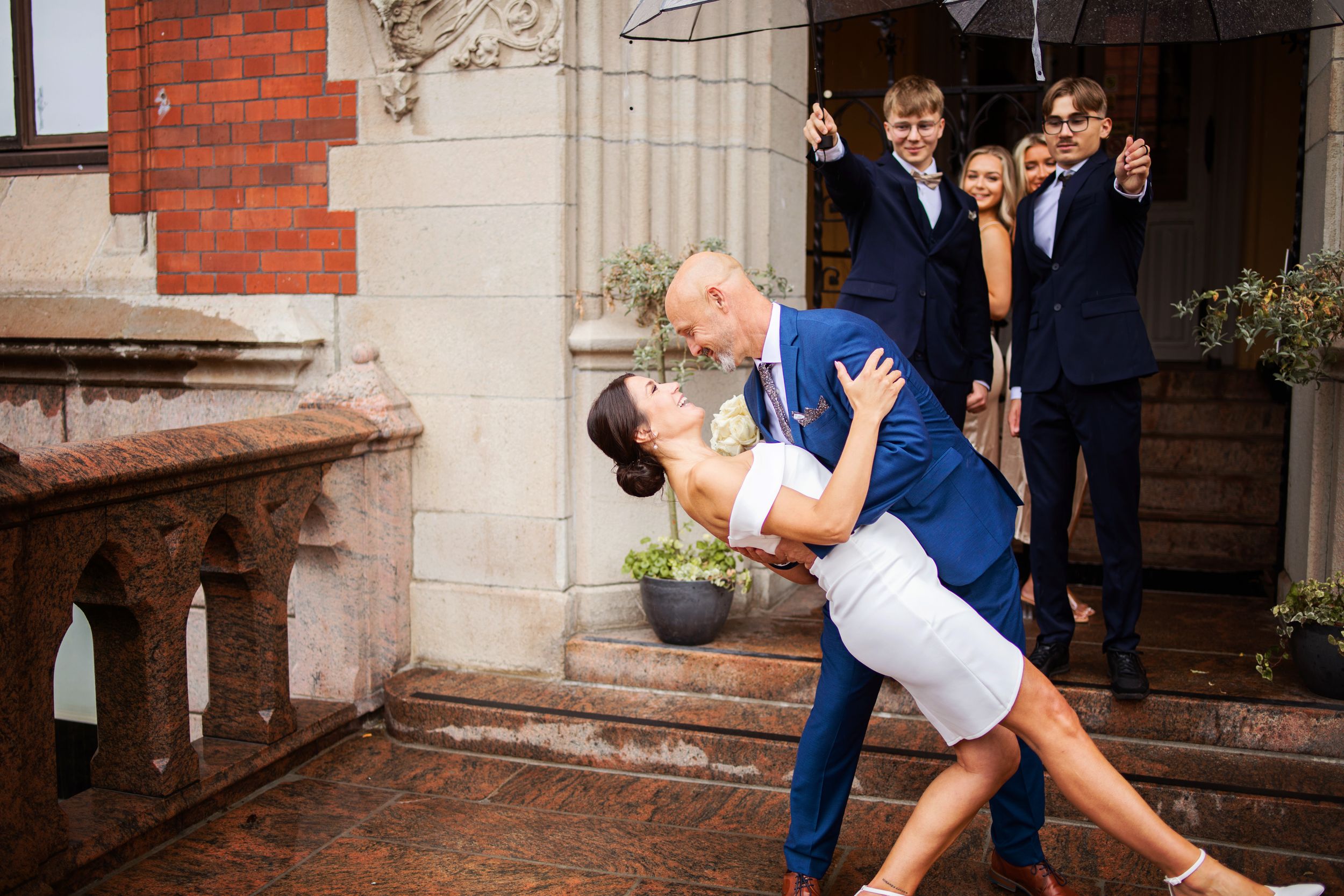 Bride and groom share romantic dip kiss outside historic building while wedding guests hold umbrellas in rain.