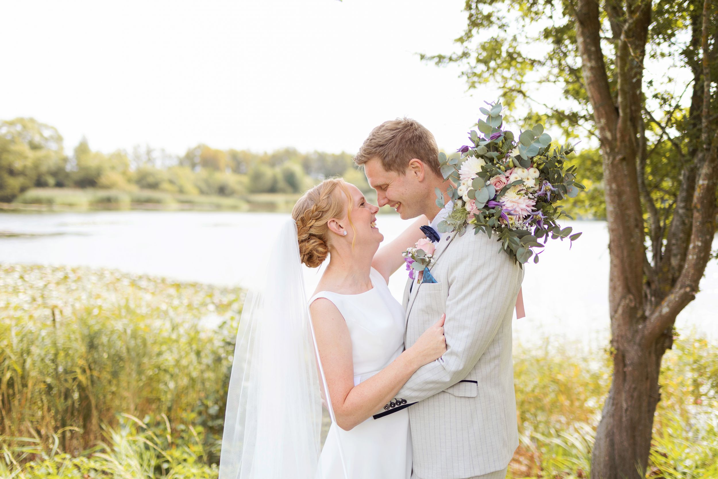 Bride and groom share a romantic moment by a lakeside, with the bride holding a purple floral bouquet.