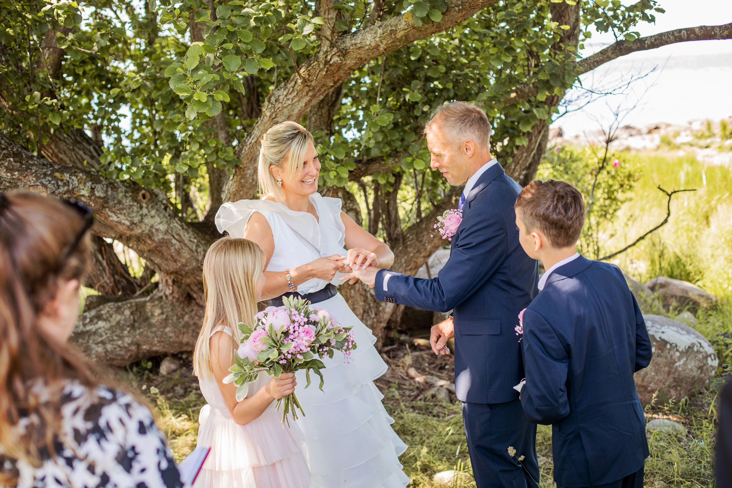 Bride and groom exchanging rings outdoors under a tree with children present at an intimate wedding ceremony.
