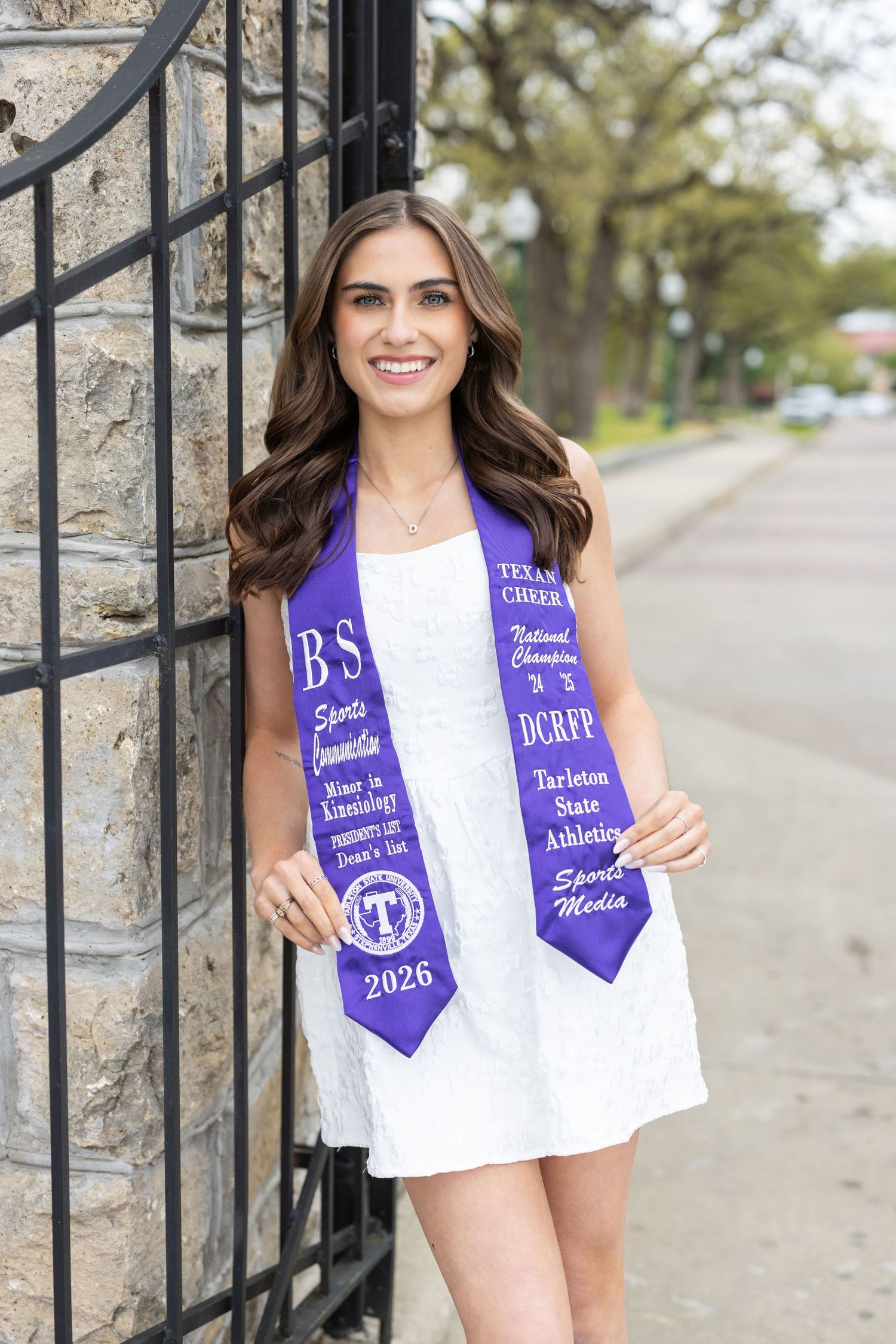 Smiling brunette woman in white dress wearing a purple Tarleton State University 2026 graduation stole outdoors.