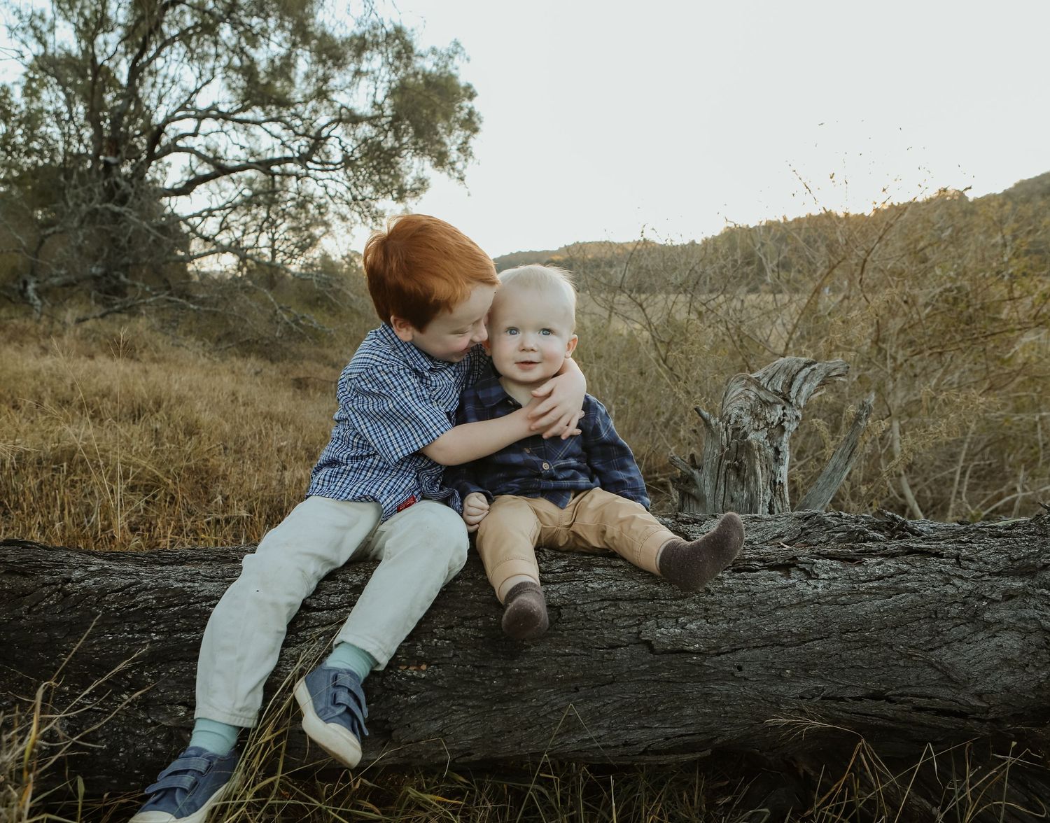 Two young siblings sit on a fallen log in a golden field, older child hugging baby affectionately outdoors.