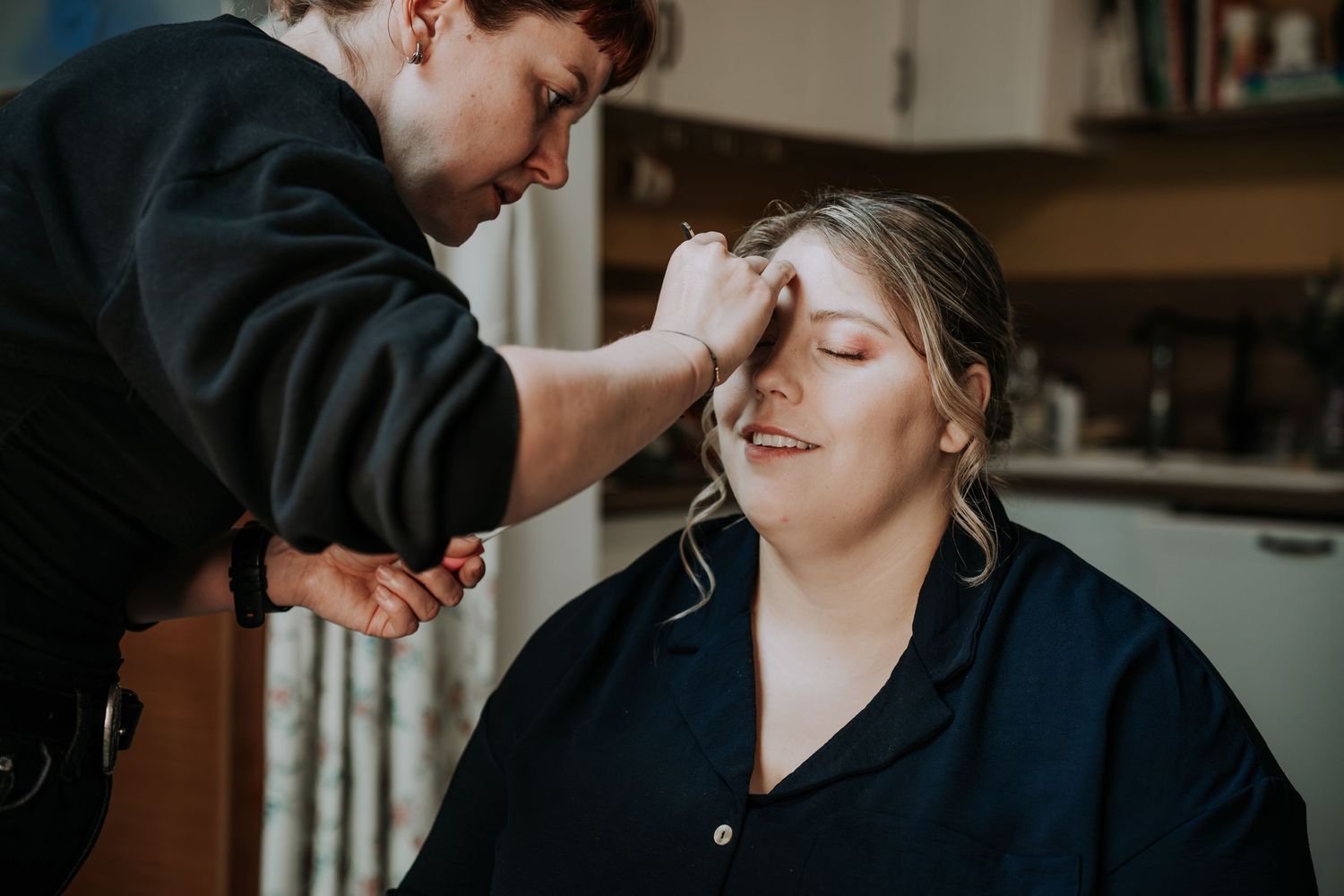 Makeup artist applying eyeshadow to a smiling woman in a navy shirt during bridal preparation.