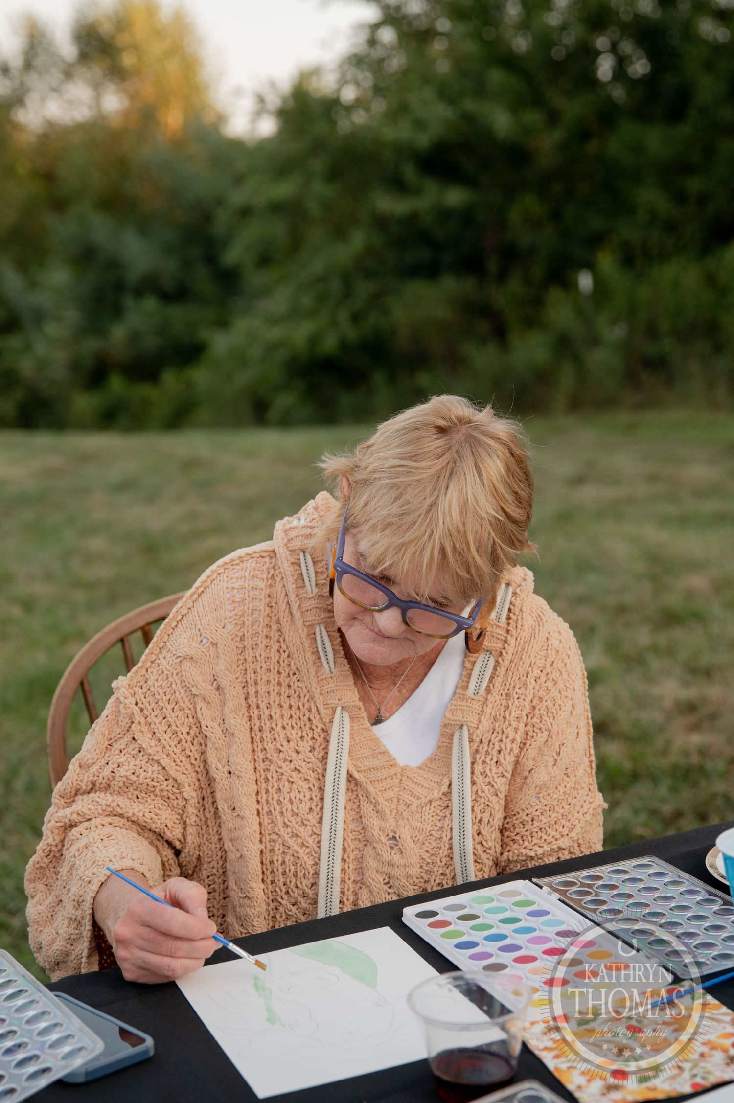 Older woman with glasses painting watercolors at outdoor table, wearing a cozy knit cardigan in a lush green field.