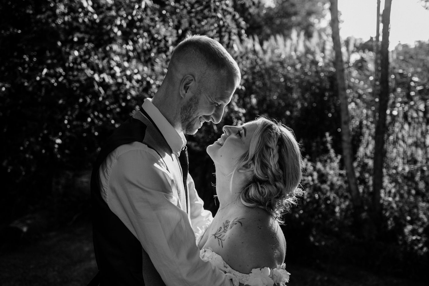 Black and white wedding photo of bride and groom sharing an intimate moment outdoors surrounded by lush greenery.