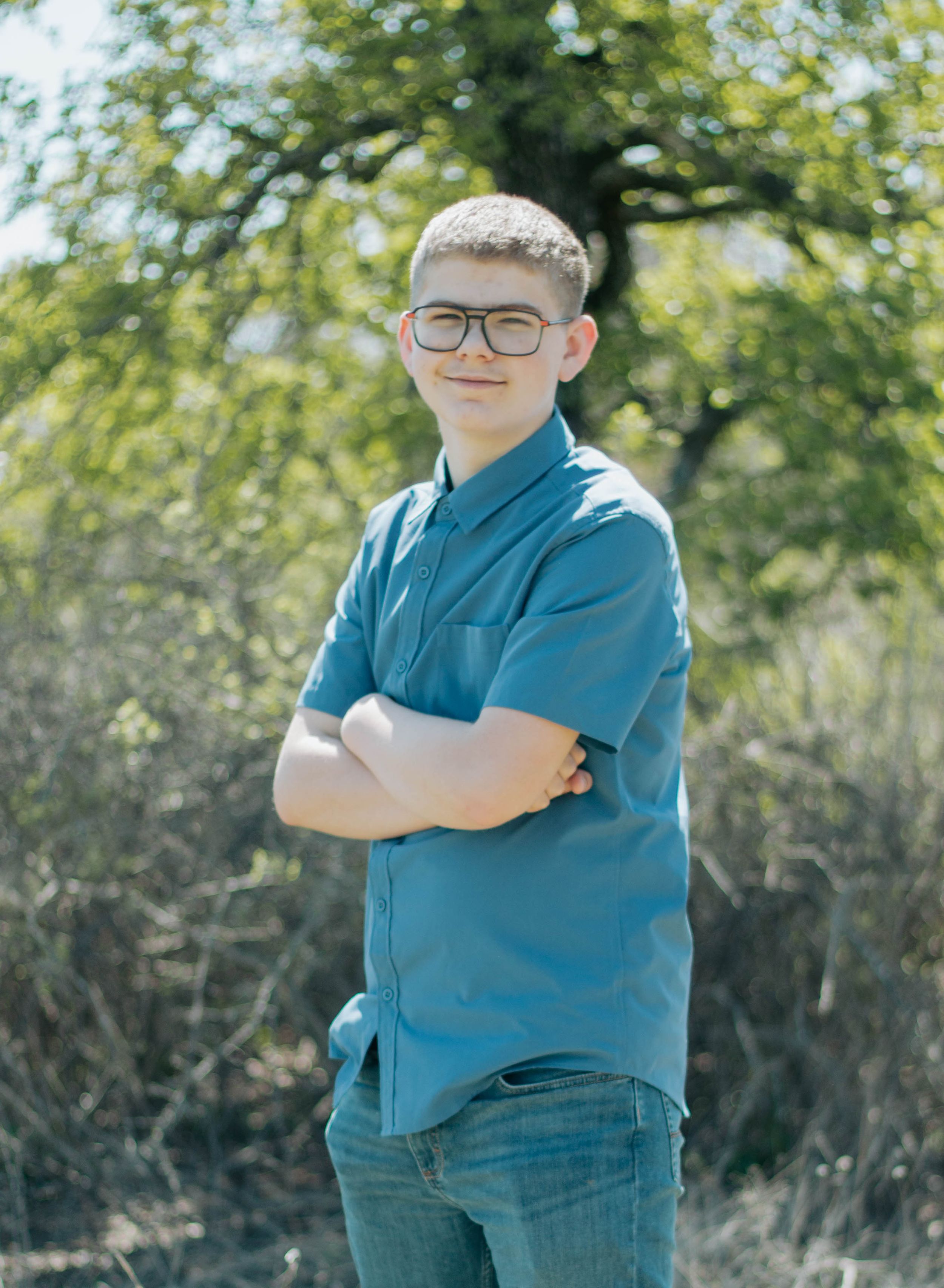 Young teen boy with glasses in teal shirt stands arms crossed outdoors near trees for a portrait photo.