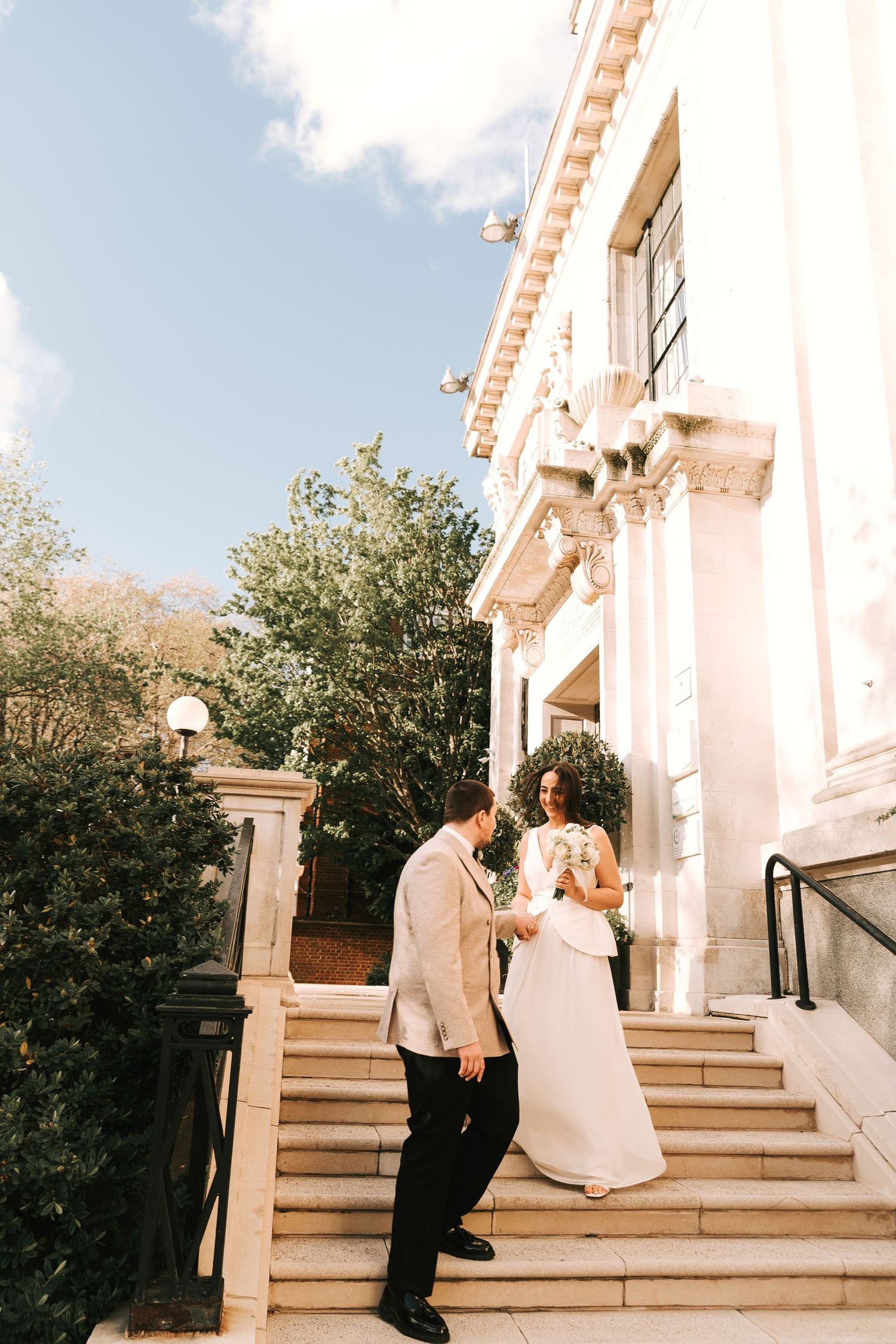 Bride and groom descend stone steps of elegant classical building, bride holding white bouquet in sunlight.