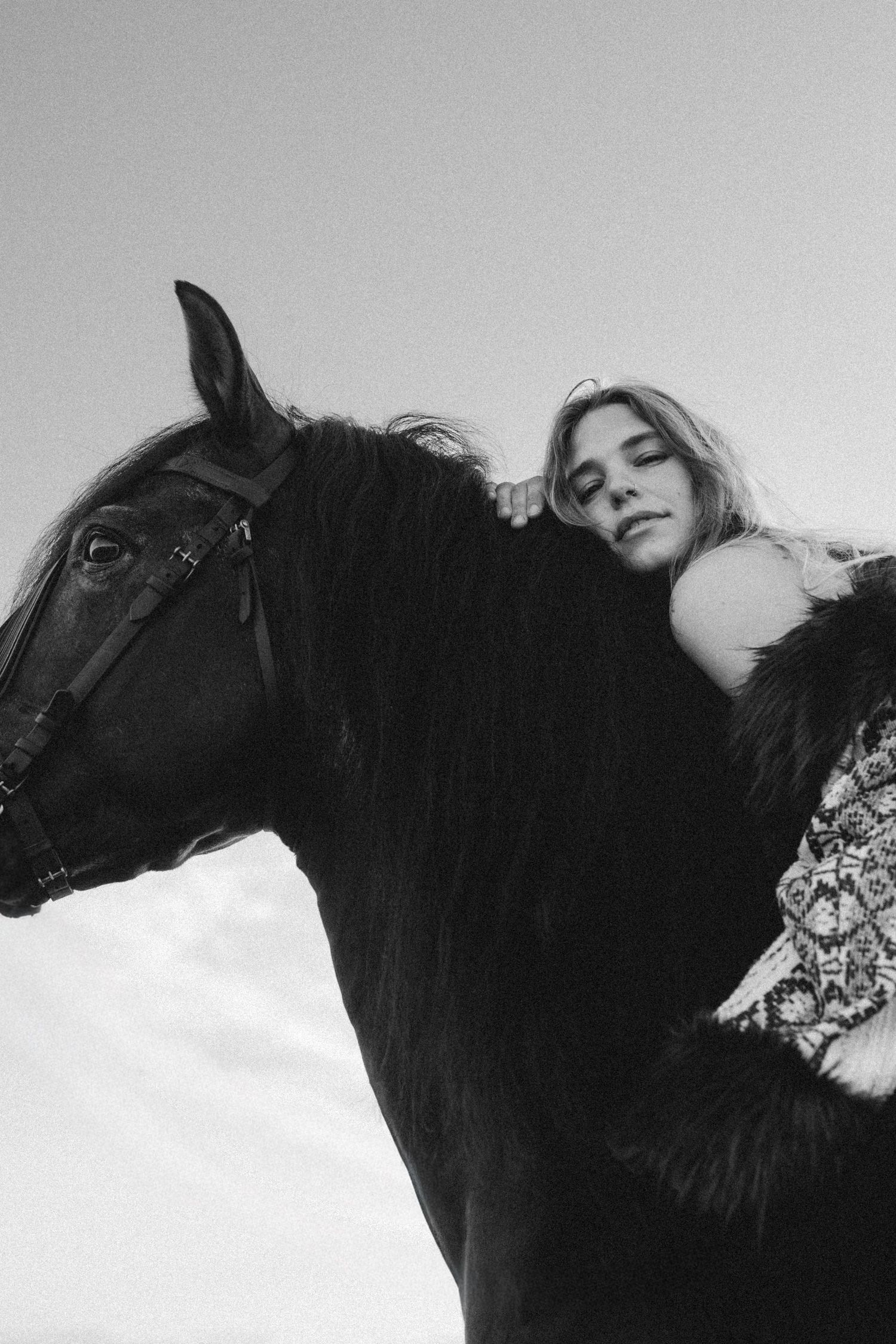 Black and white photo of a young woman resting her head on a dark horse, against a cloudy sky. Javea photographer