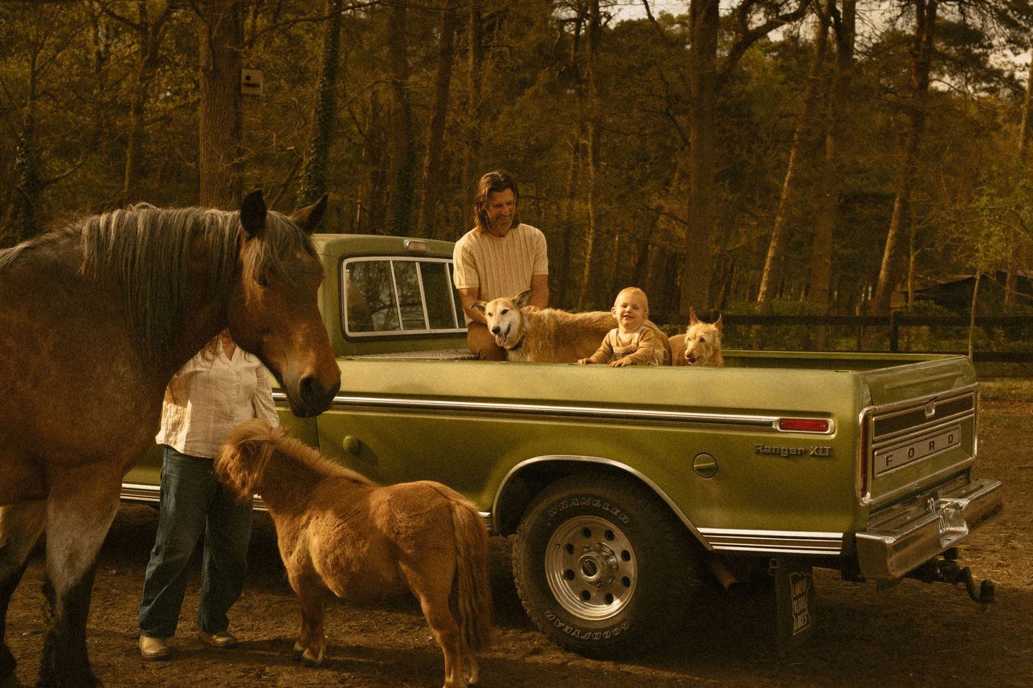 A family with a horse, pony, dogs, and child gathered around a vintage green pickup truck in a wooded setting. Javea photographer