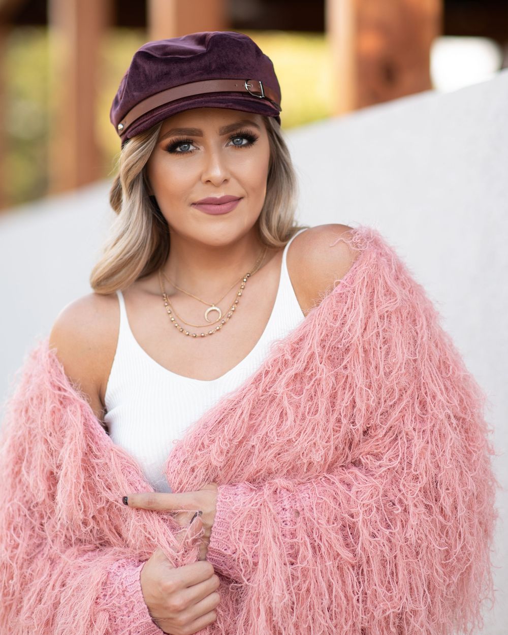 Woman in pink fluffy coat and purple baker boy hat poses confidently outdoors.
