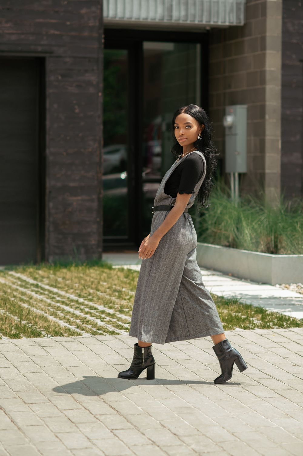 A stylish woman in gray culottes, black top, and ankle boots walks confidently on a paved path outside a modern building.