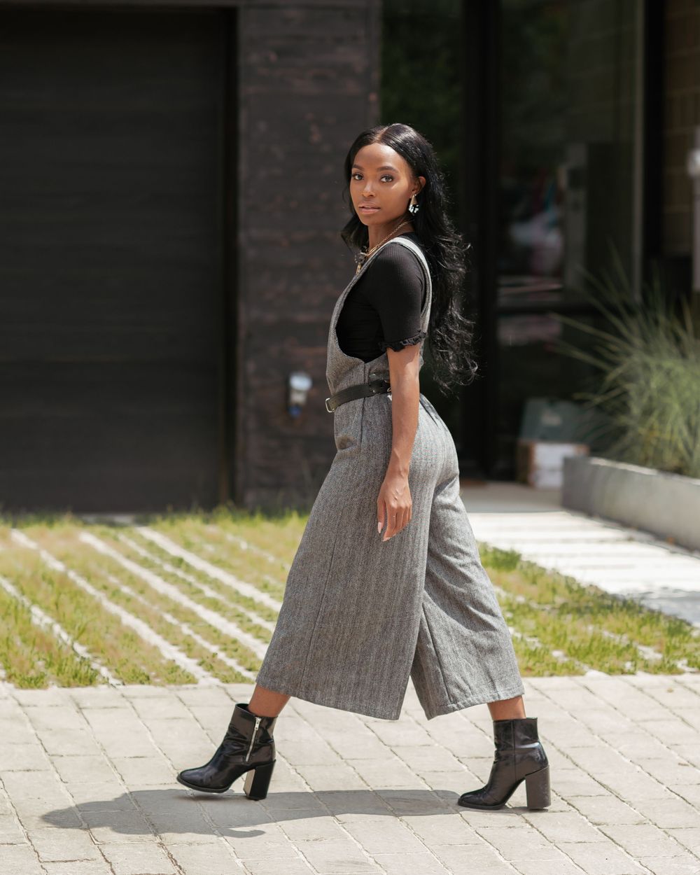 Black woman in gray linen overalls, black top, and ankle boots walking on a brick pathway outdoors.