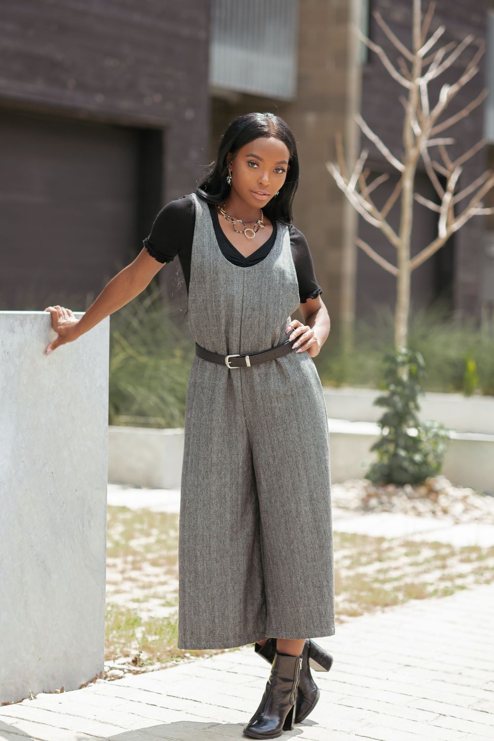Model wearing gray herringbone wide-leg jumpsuit with black top and leather belt, posing outdoors in black heeled boots.