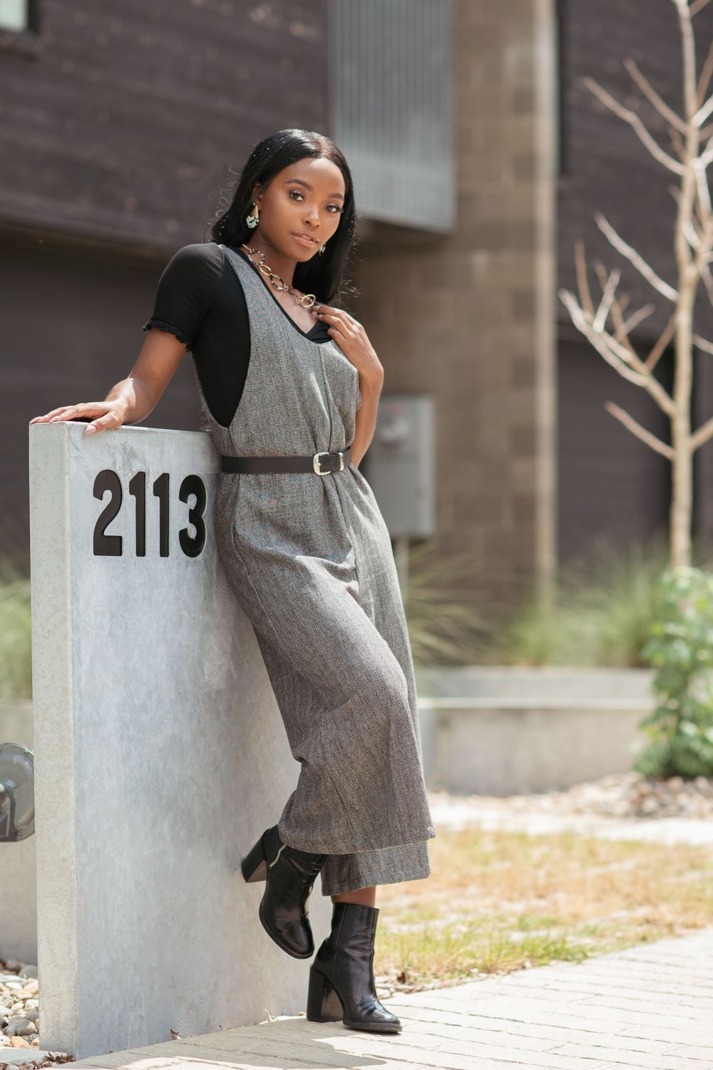 Stylish woman in gray belted jumpsuit and black boots leaning against a concrete post numbered 2113.
