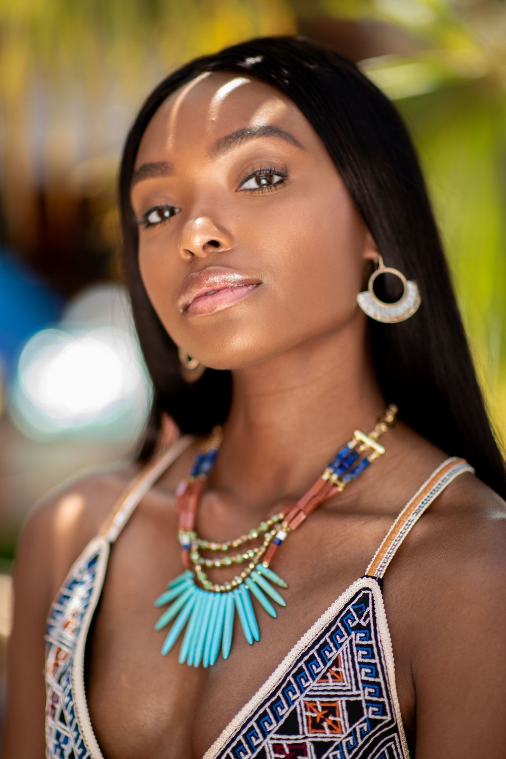 Young Black woman wearing turquoise statement necklace and hoop earrings in colorful tribal print dress outdoors.