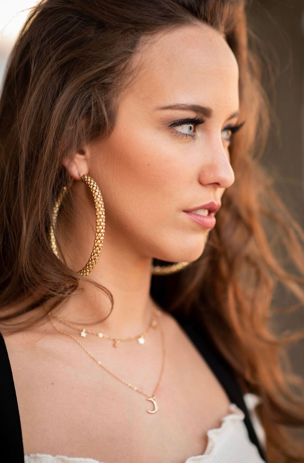 Young woman wearing gold hoop earrings, layered necklaces with moon pendant, and white top, side profile view.