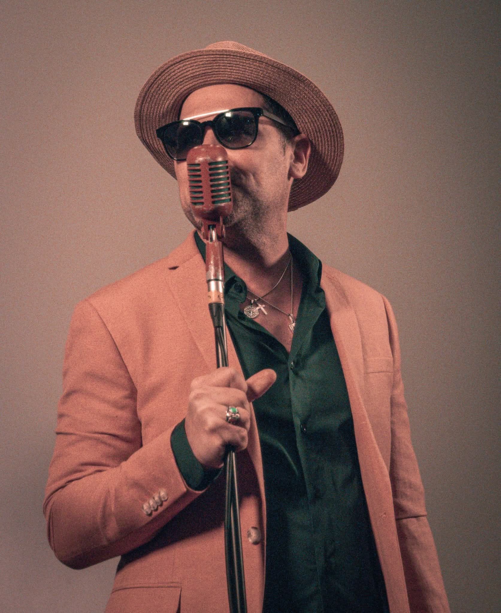 Male singer in pink blazer and fedora holding vintage microphone on stage.