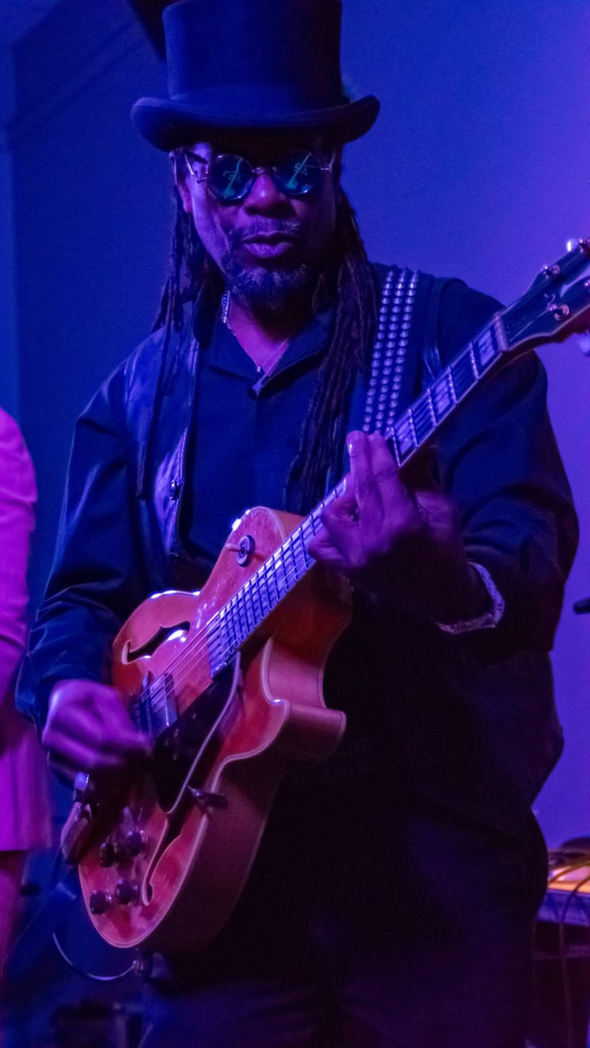 Guitarist in top hat and dreadlocks playing hollow-body electric guitar under purple stage lights.