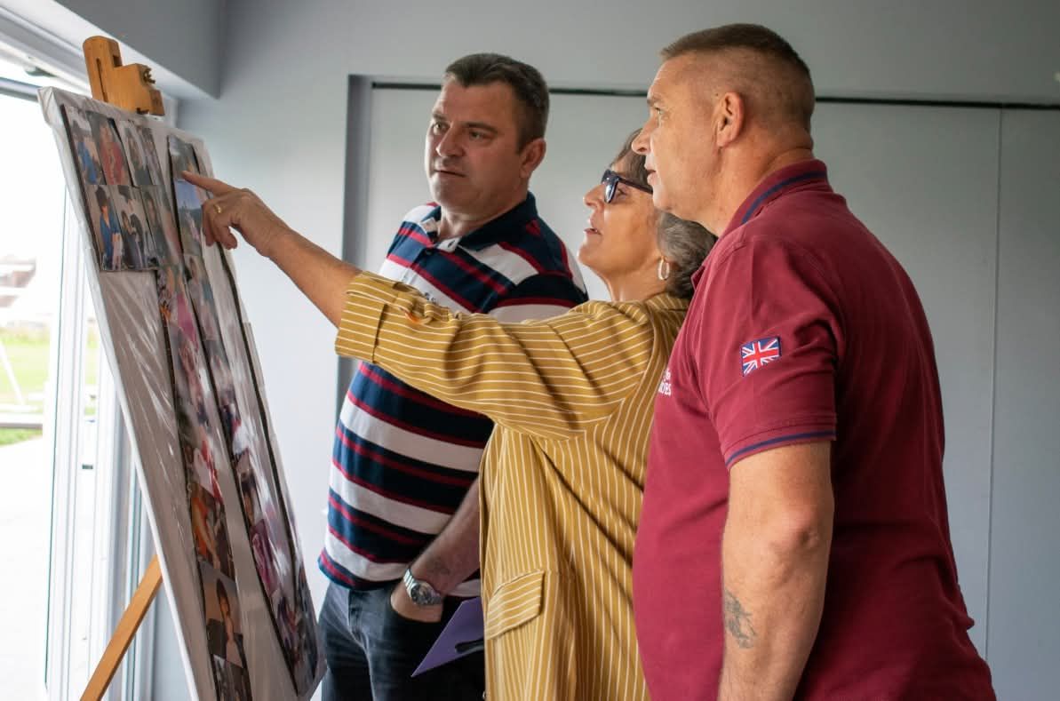 Three people examining a photo collage display board at an indoor event or exhibition.