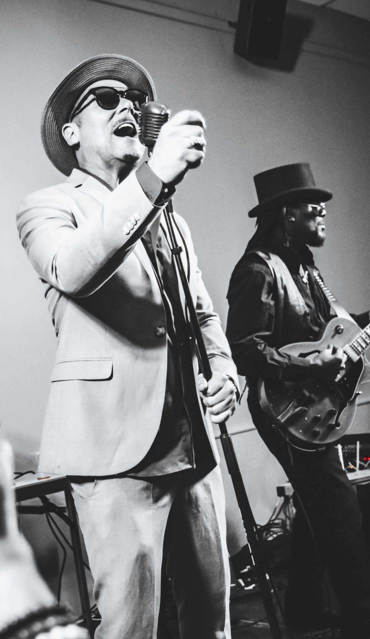 Black and white photo of male vocalist singing with guitarist in top hat performing live.