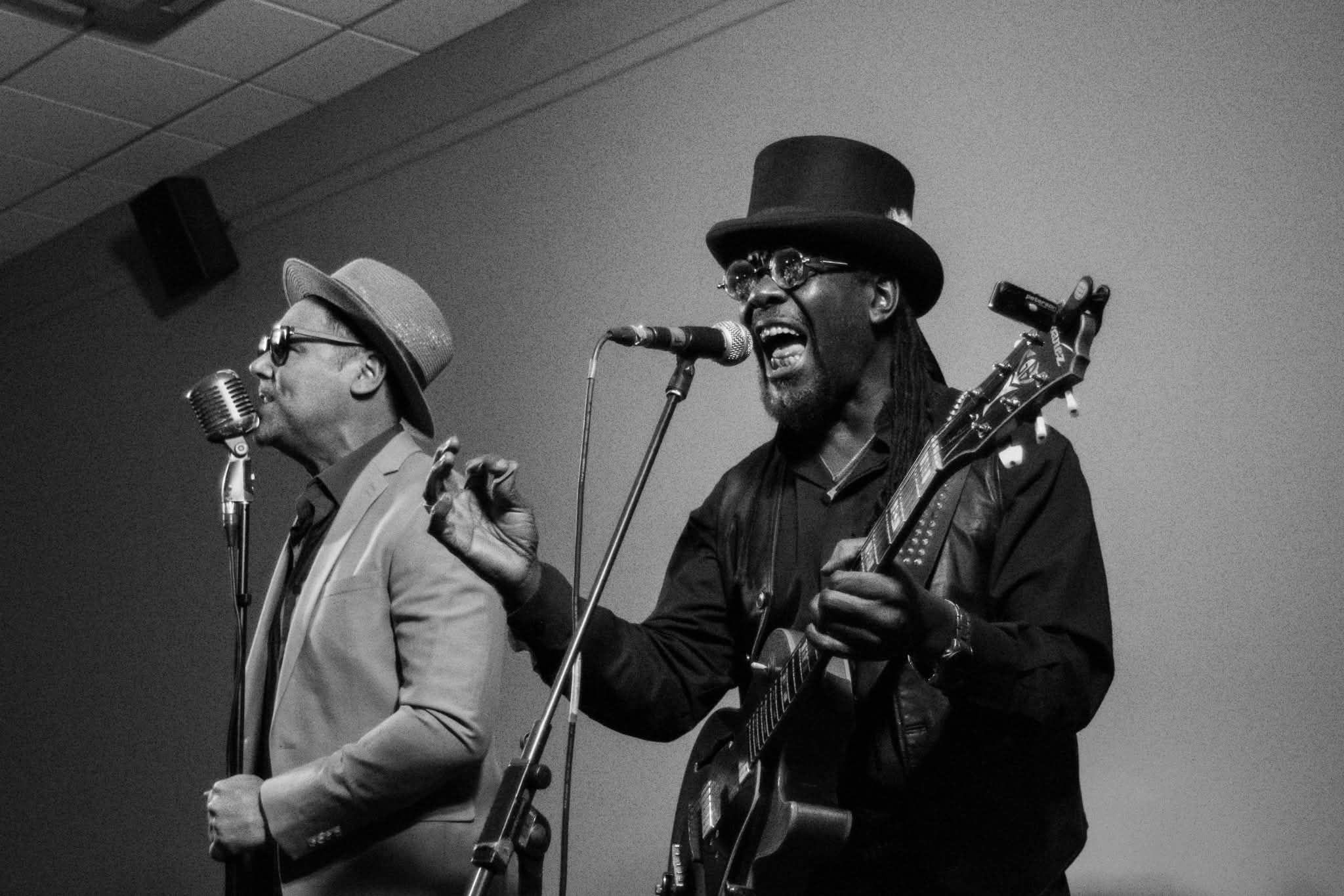 Two male musicians performing live, one singing and one playing guitar in black and white.