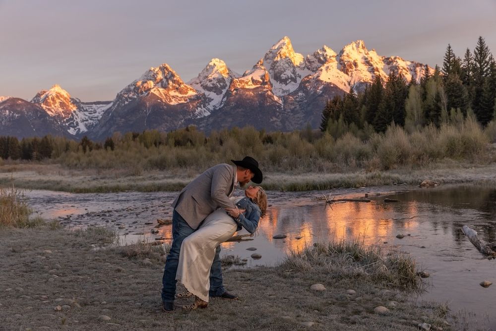 Couple sharing a romantic dip kiss at sunset with Grand Teton mountains reflected in calm water behind them.