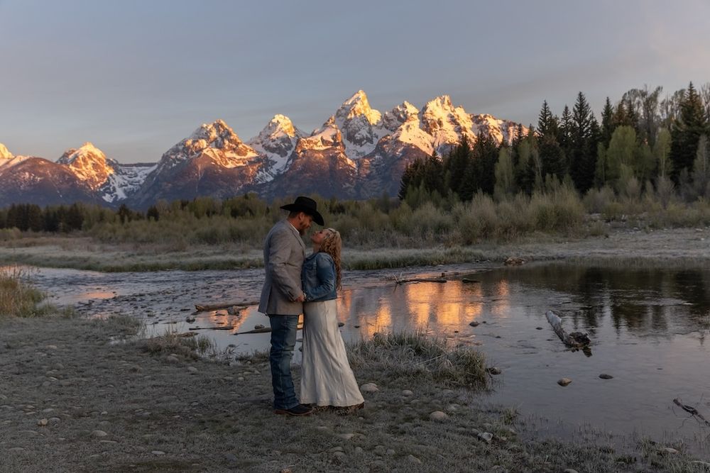Couple embracing at sunset with snow-capped Grand Teton mountains reflected in a calm river, surrounded by autumn trees.