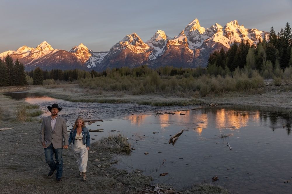 Couple walking along a river at sunset with the snow-capped Grand Teton mountains reflecting in the water.