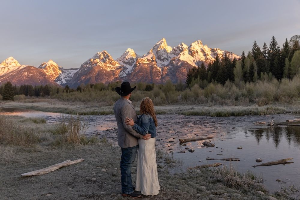 Couple embraces at sunset by a river with snow-capped Grand Teton mountains behind them.