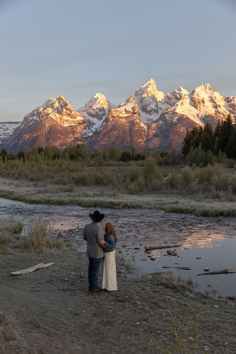 Couple in western attire stands by rocky river with stunning Grand Teton mountain sunset view.