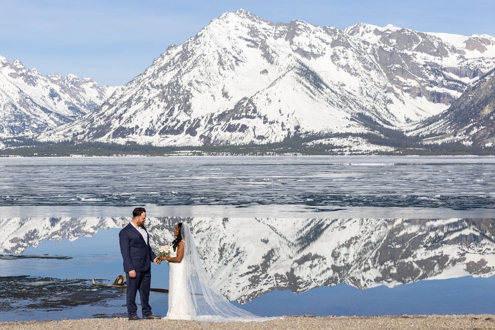 Bride and groom stand by a reflective mountain lake with snow-capped Grand Teton peaks in the background.