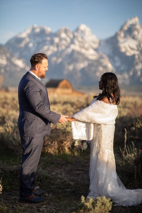 Couple exchanges vows at Grand Teton National Park wedding, bride in lace gown with fur wrap, groom in gray suit.