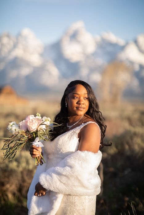 Bride in white lace gown and fur wrap holds pink bouquet with snow-capped Grand Teton mountains behind her.