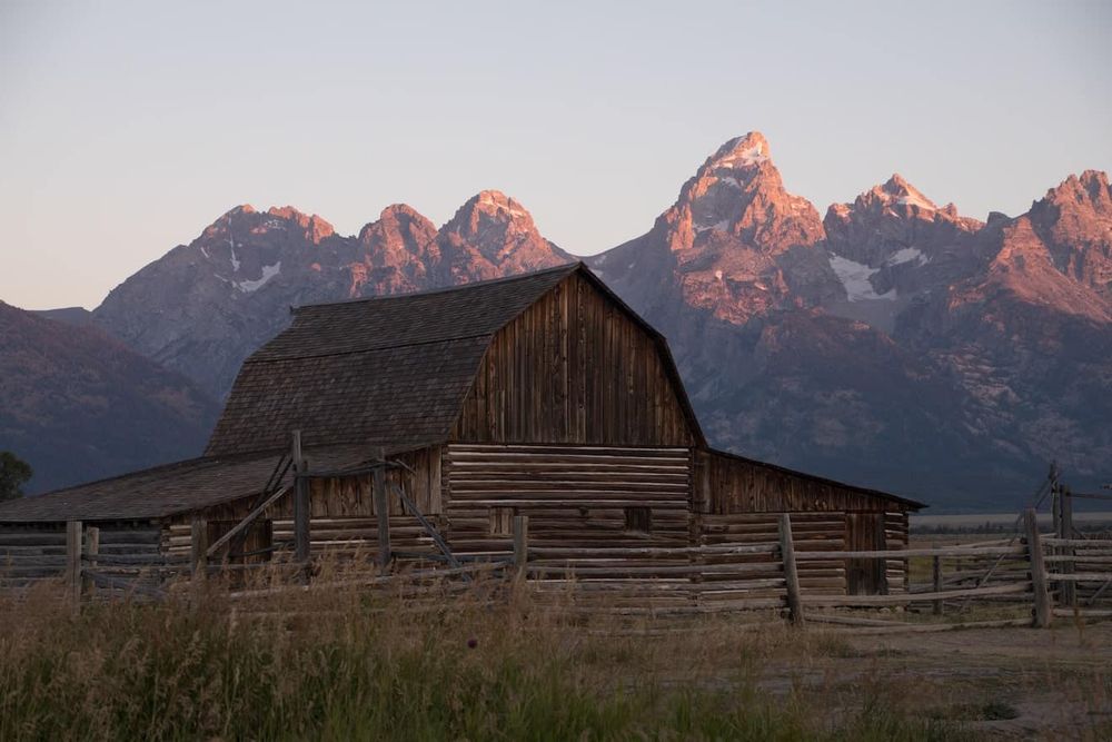 Historic wooden barn at sunset with snow-capped Grand Teton mountains glowing pink in the background.