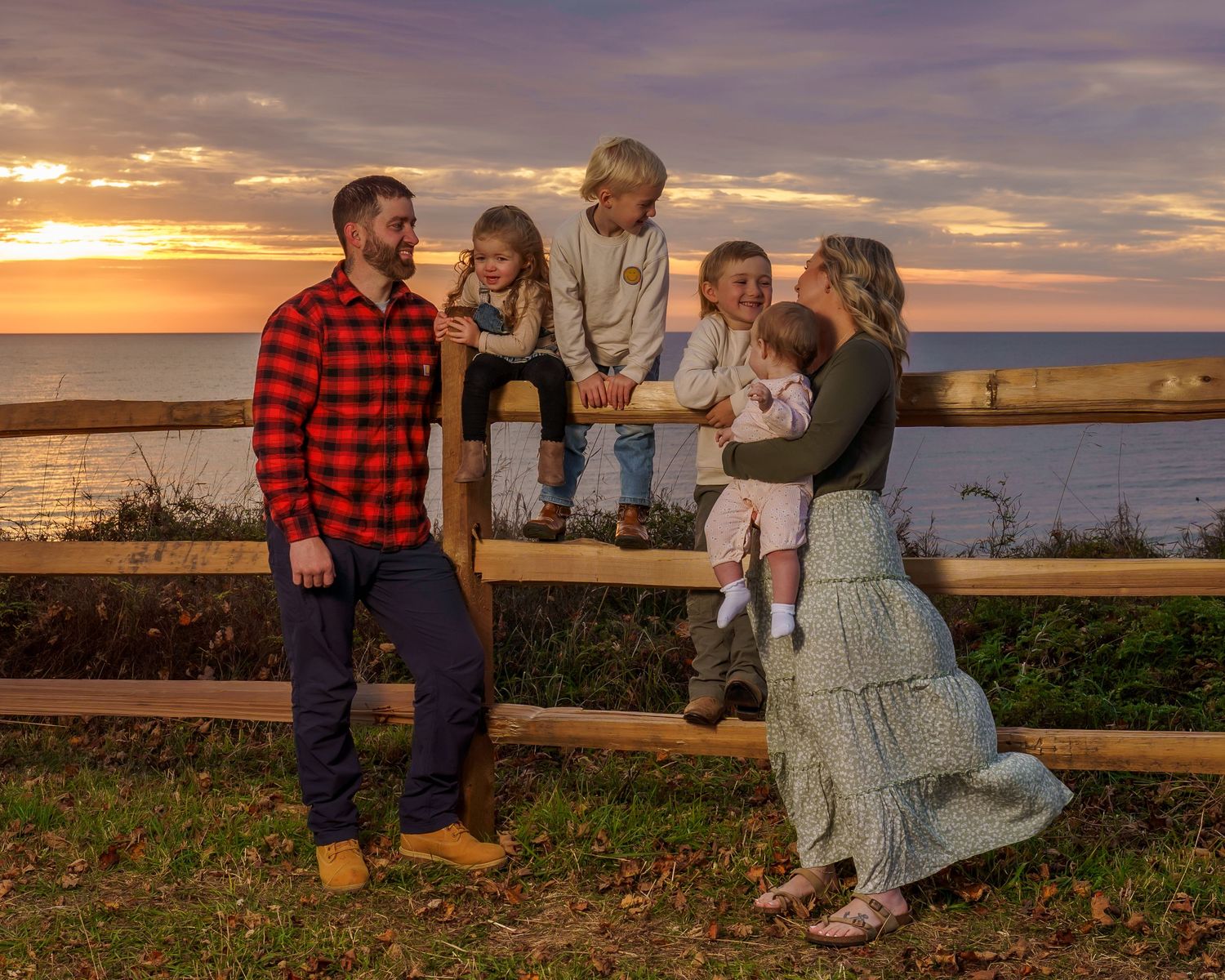 Family of six poses at a wooden fence during a stunning ocean sunset, wearing casual fall outfits in warm tones.