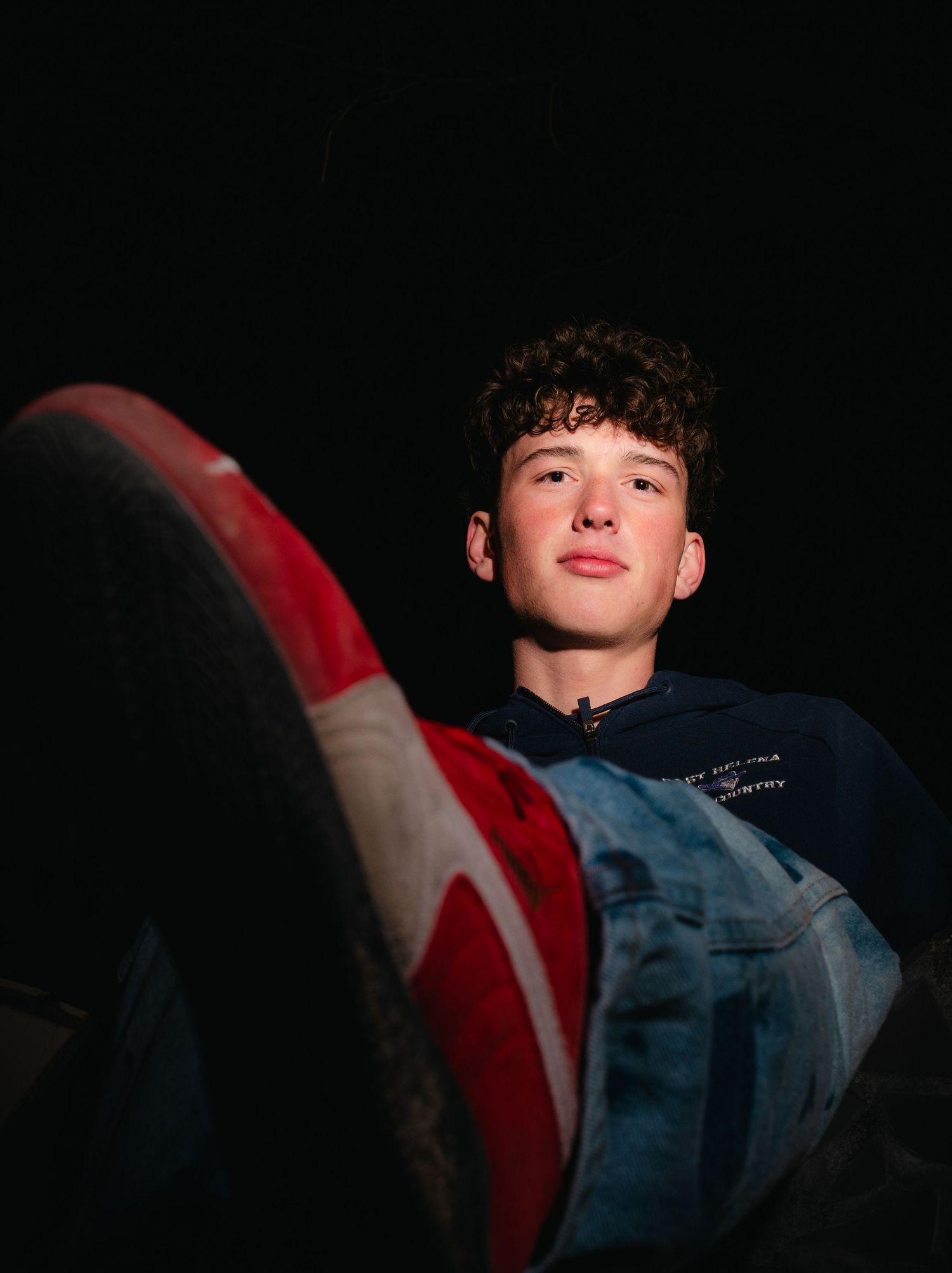 Young man in navy hoodie and jeans relaxing, shot from low angle with red and white sneaker sole in foreground.