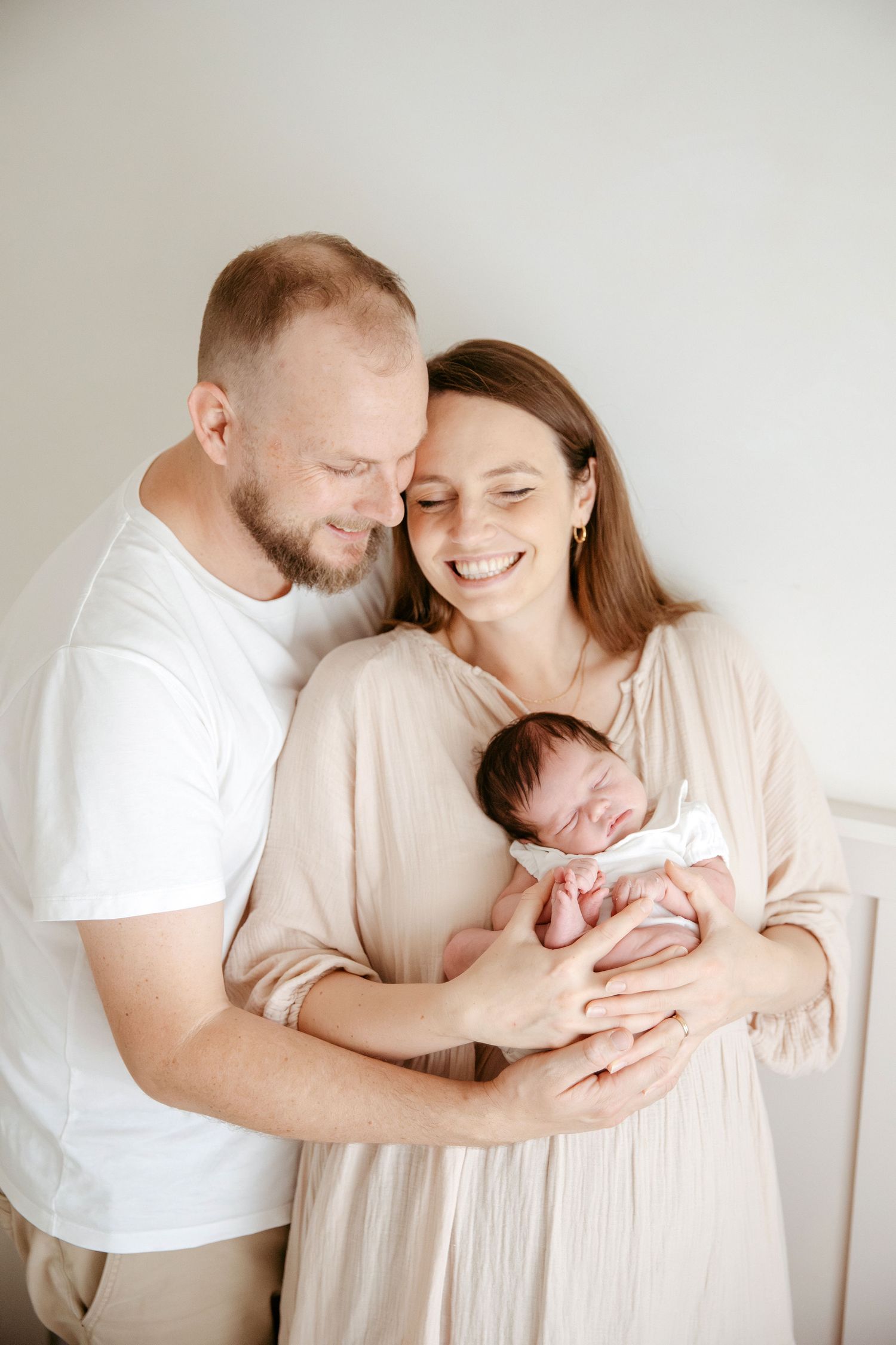 Happy parents smiling and holding their sleeping newborn baby, dressed in neutral tones against a white background.