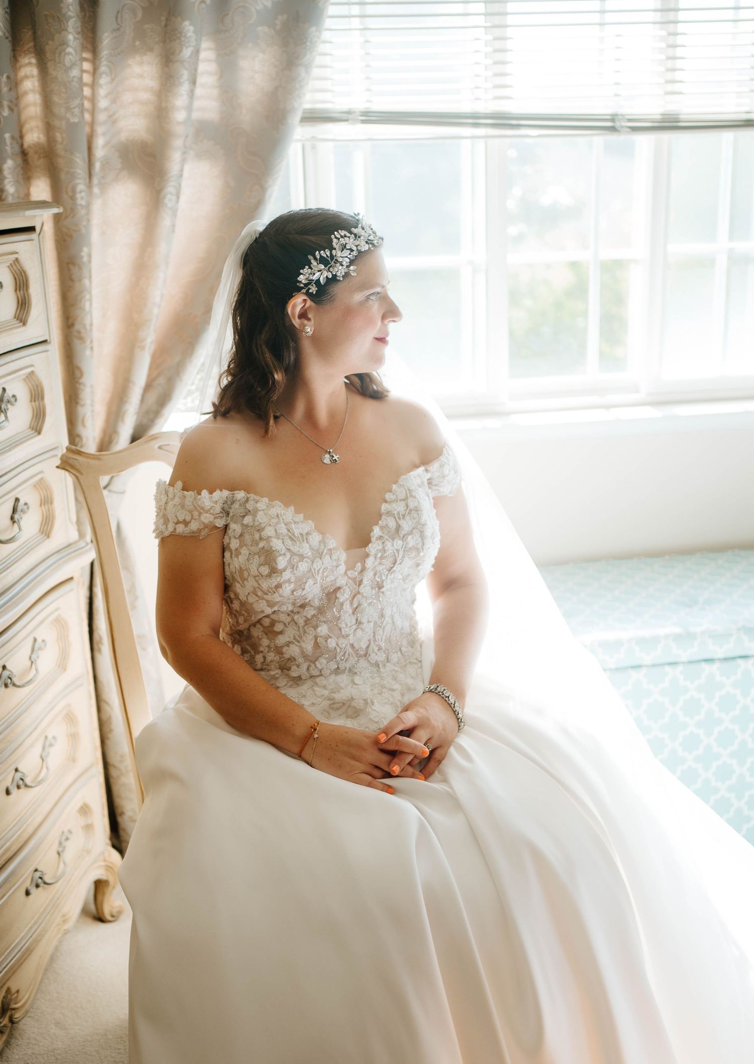 Bride in off-shoulder lace wedding gown with floral headpiece sits by window in soft natural light.