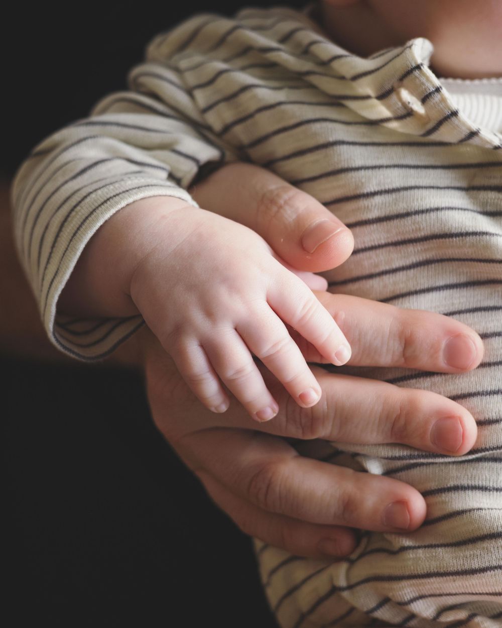 A baby's tiny hand resting gently on an adult's hand, both wearing striped clothing, symbolizing parental love.
