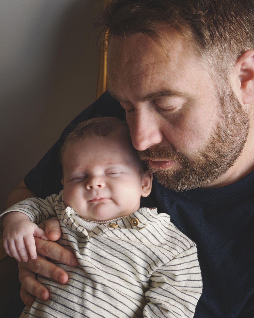 A bearded father tenderly holds his sleeping newborn baby, both with eyes closed, sharing a peaceful bonding moment.
