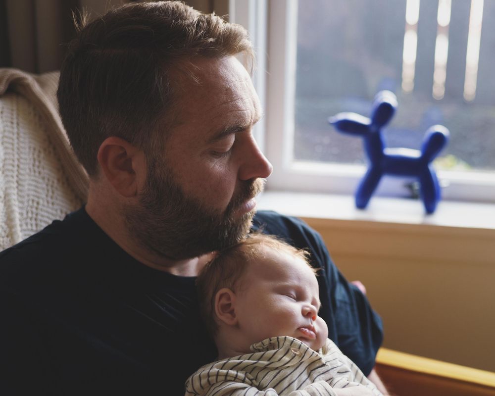 A bearded father tenderly holds his sleeping newborn baby near a window with a blue dog figurine in background.