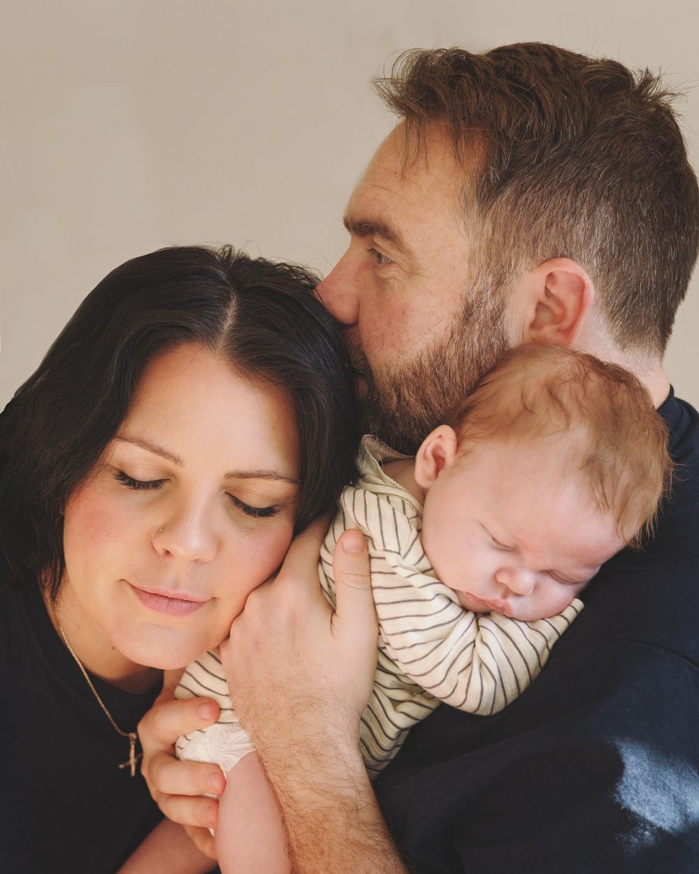 A bearded man kisses a dark-haired woman holding a sleeping baby in a striped onesie in a tender family portrait.