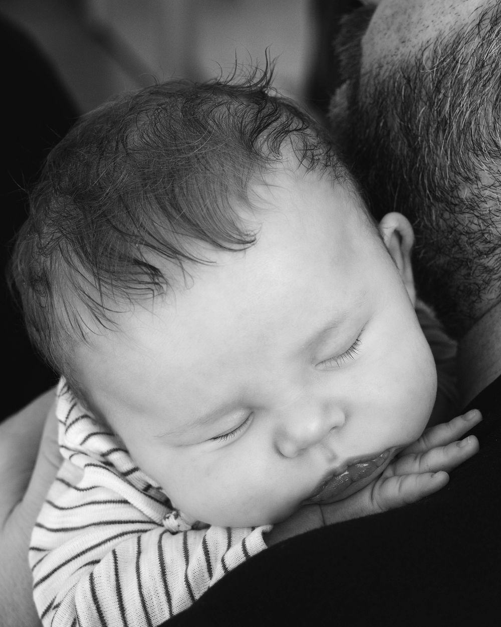 Black and white photo of a sleeping newborn baby with dark hair resting peacefully on an adult's shoulder.