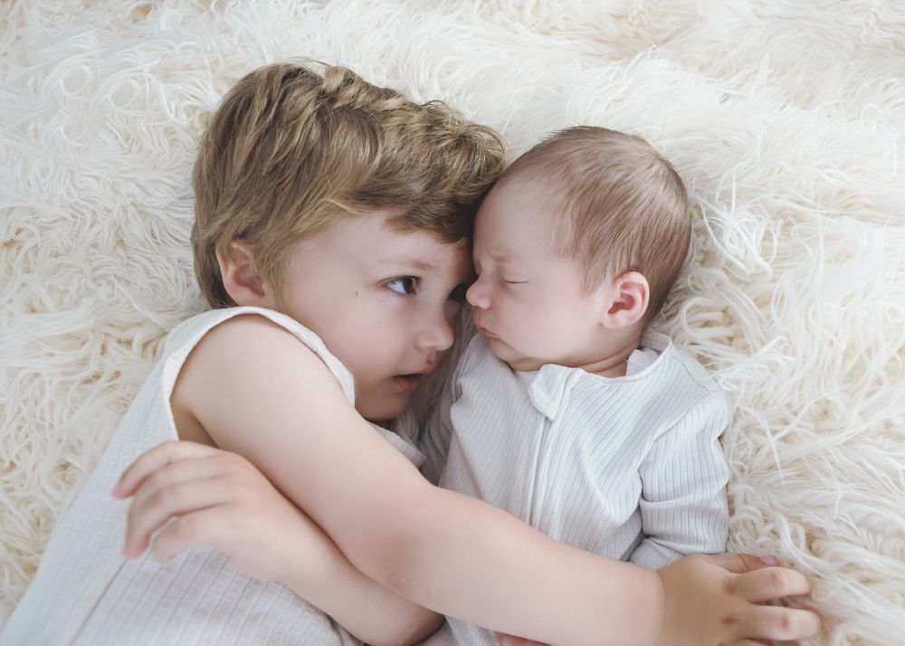 Toddler gently hugging a sleeping newborn baby on a soft white fluffy blanket.